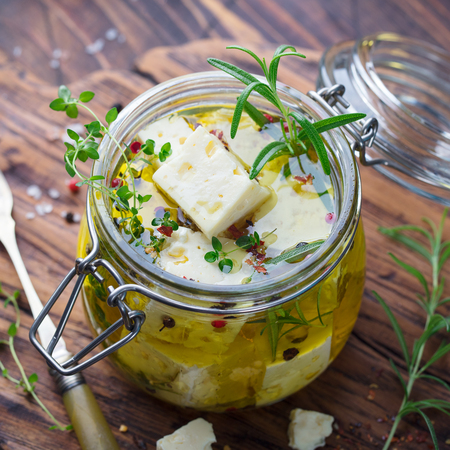 Feta cheese marinated in olive oil with fresh herbs in glass jar. Wooden background. Close up.の写真素材