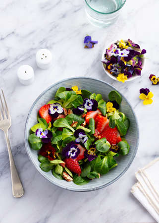 Fresh green salad with strawberries and edible flowers in a bowl. Marble background. Top view.の写真素材