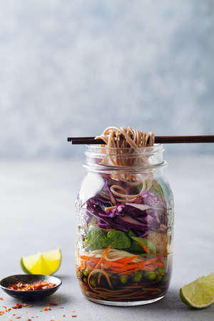 Healthy asian salad with noodles, vegetables, chicken and tofu in glass jars. Grey background. Copy...