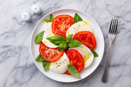 Caprese salad with tomatoes, mozzarella cheese and basil on a white plate. Marble background. Close up. Top view.の写真素材