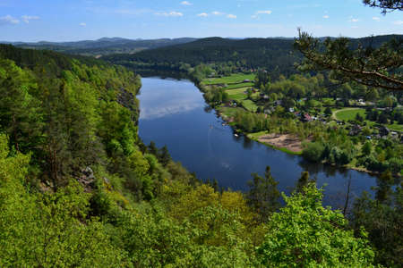 Beautiful village in woods along Vltava river. Lush green trees, yellow fields, blue sky, white...