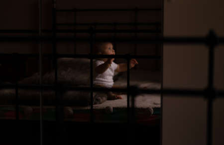 Infant boy sits on bed in dark room. Holds toy in hands. Black negative space. High quality photoの写真素材