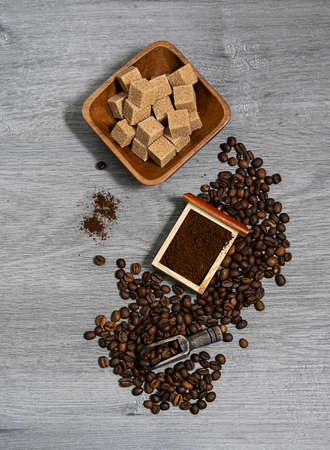 Coffee beans are scattered on a light wooden table. A box of ground coffee and pieces of cane sugar in a wooden bowl in the background. Top viewの写真素材