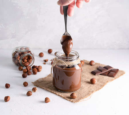 Chocolate-nut paste in a glass jar on a light table. With a spoon, the paste pours into the jarの写真素材