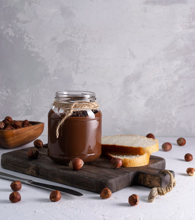Chocolate-nut paste in a transparent jar on a dark brown wooden board. Hazelnuts are scattered on a light table. Pieces of white bread are lying on a board. Nuts in a wooden bowl in the backgroundの写真素材
