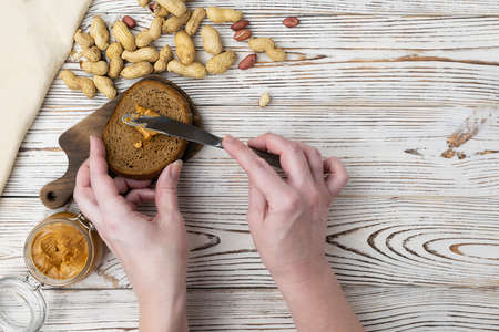 Hands spread peanut butter on bread. Pasta in a glass jar on a light wooden table. Peanuts are...