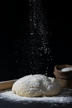 Dough, rolling pin and a canvas bag of flour on a dark table