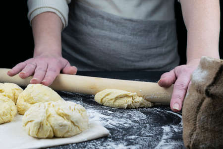 Hands roll out the dough. Ingredients for making bread and tortillas. Pieces of dough and flour in a canvas bag in the background. Dark background. The table is strewn with flourの写真素材