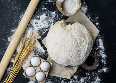 Dough sprinkled with flour, rolling pin and ears of wheat on a black table. Raw eggs, flour in a canvas bag in the background. The table is strewn with flour. top viewの写真素材