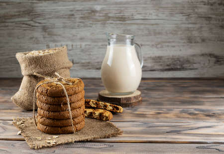 Oatmeal biscuits in a stack tied with canvas rope on a light-screaming wooden table. Oatmeal in a canvas bag and a jug of milk in the backgroundの写真素材
