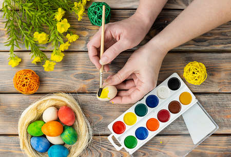 Colorful eggs in a wicker basket, paints and herbs on a light brown wooden background. Hands hold a brush and paint a wooden egg. Preparing for Easterの写真素材