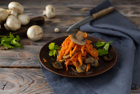 Korean carrot salad with fried mushrooms and cilantro. Raw mushrooms on a wooden plank in the background. Salad in a clay plate with greensの写真素材