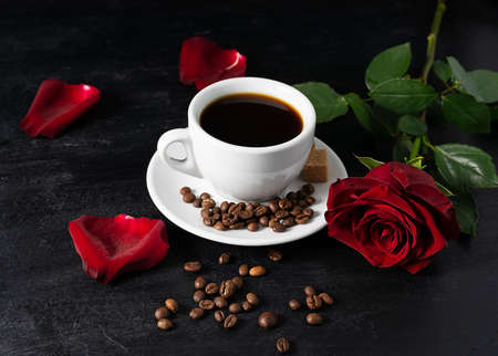 Black coffee in a white cup on a black table. A red rose and petals in the background. Coffee beans on the tableの写真素材