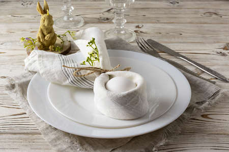 An Easter egg in a light napkin on a white plate. A candle in the form of a rabbit and wine glasses...