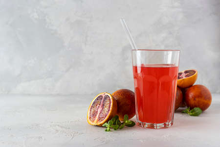A tall glass of red orange juice on a light table. Red oranges and mint leaves in the background....