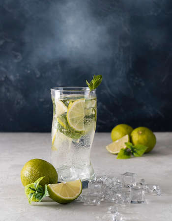 A mojito cocktail with ice in a glass glass on a light table. Slices of lime, sprigs of mint and ice cubes in the background. Blue background, space for textの写真素材