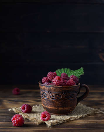 Large ripe raspberries in a clay cup on a canvas napkin. Dark brown wooden background