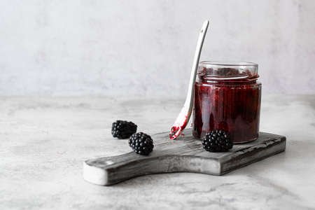 A glass jar with blackberry jam on a light wooden plank. Large blackberries on a marble table. A...