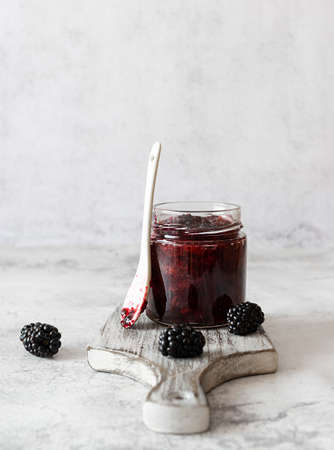 A jar of blackberry jam on a light wooden plank. Large berries on the table. A white ceramic spoon in the background. light backgroundの写真素材