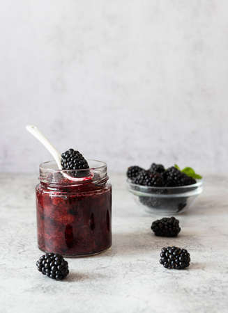 Blackberry jam in a transparent jar on a light table. Ripe blackberries in a glass bowl in the...