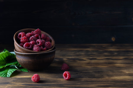 Clay bowls with ripe raspberries on a dark wooden table. Raspberry leaves in the background. Dark...
