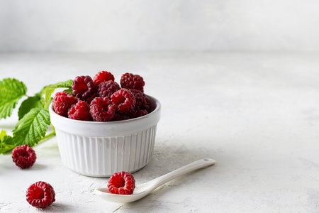 Ripe juicy raspberries in a white ceramic vase on a light table. green leaves in the background. A...