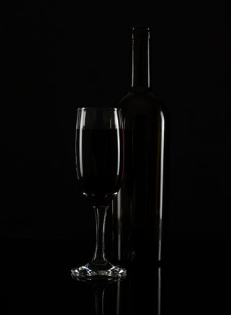 A tall glass of red wine and a bottle on a black table. White highlights on the glass and bottle. black backgroundの写真素材