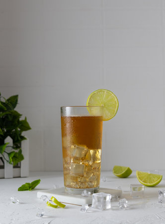 Iced tea with ice and lime in a tall transparent glass. Ice cubes and lime wedges in the background. Sprigs of mint in a basket on the table. light backgroundの写真素材