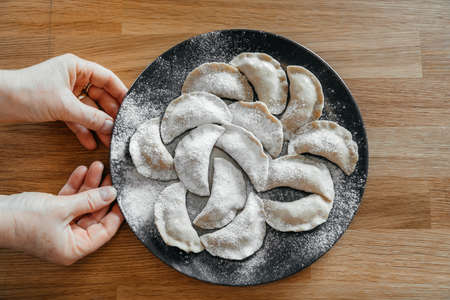 Stuffed homemade dumplings on a dark plate with flour on wooden table with cook's hands. Ukrainian vareniki. Close up. Top view.の写真素材