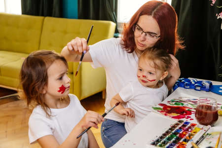 Family hobbies leisure activity home concept. Mother and small daughter drawing with fun using paintbrushes and watercolors. Children and parent creating picture together, body paint, home quarantine.の写真素材