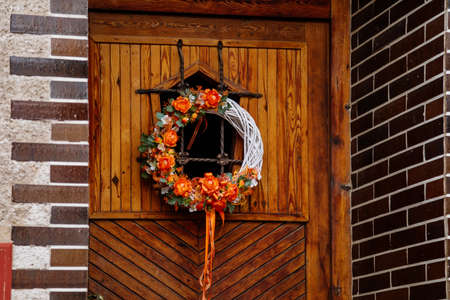 Handmade wreath decorated by red, yellow and orange flowers, green leaves and festive ribbons, symbol of happiness and prosperity, easter decoration on vintage antique wooden door, red brick house.の素材 [FY310171629427]