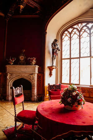 Lednice, Southern Moravia, Czech Republic, 04 July 2021: Castle interior with neo-gothic wooden carved ceiling and furniture, red salon, table with chairs and sofa, open marble fireplace, chandelier.のeditorial素材