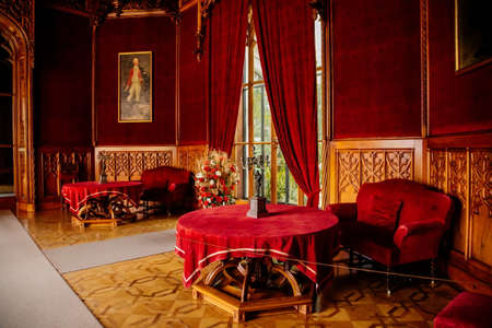 Lednice, Southern Moravia, Czech Republic, 04 July 2021: Castle interior with neo-gothic wooden carved ceiling and furniture, red salon, table with chairs and sofa, open marble fireplace, chandelier.のeditorial素材