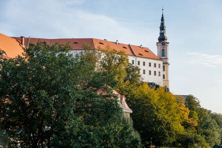 Decin, North Bohemia, Czech Republic, 2 October 2021: castle on cliff above river Elbe, white tower with pointed roof, trees near mountain on the embankment, medieval town at autumn sunny day.のeditorial素材