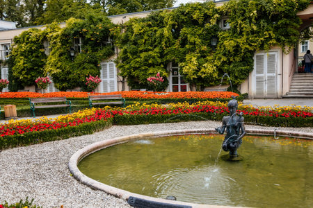 Salzburg, Austria, 28 August 2021: Papagenabrunnen or Papagena fountain by sculptor Josef Magnus in Orangerie of Mirabell gardens, bronze statue of girl with bird, palace at summer day, Red flowersのeditorial素材