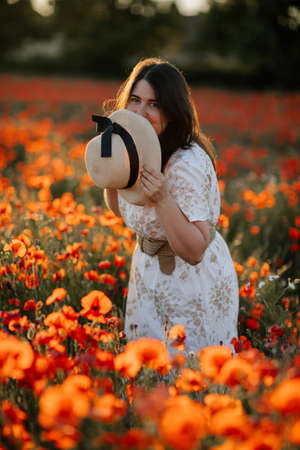 Scenic picturesque of beautiful girl in white and beige dress and straw hat in poppy field at sunset, Portrait of romantic young woman with flowers, body positivity, Selective focus natural lightの写真素材