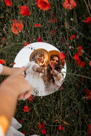 Beautiful young woman with girl in field with poppies, mother and daughter in white dresses and straw hats in evening at sunset, Summer countryside flowers, Rural simple life, reflection in mirrorの写真素材