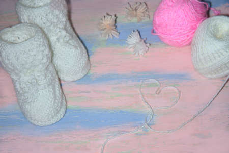 White knitted baby shoes booties on a pink-blue wooden table with balls of thread and a heart of threads.の写真素材