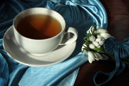 A white cup with green tea on a saucer and a small bouquet of spring white snowdrops, tied with a ribbon.の写真素材
