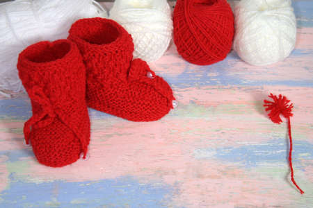 Red knitted baby booties, a red balls of wool yarn for knitting and a red pompon of yarn on a pink - blue backgroundの写真素材