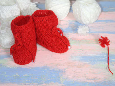Red knitted baby booties, a red balls of wool yarn for knitting and a red pompon of yarn on a pink - blue backgroundの写真素材