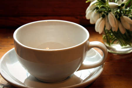 White cup for tea on a white saucer on a wooden table and a bouquet of snowdrops in the cornerの写真素材