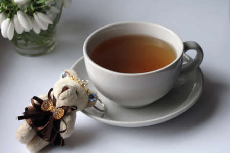 White cup with tea on a saucer on a wooden table, a key ring a bear cub and a bouquet of snowdropsの写真素材