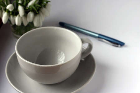 White cup for tea on a white saucer on a wooden table and a pen bouquet of snowdrops in the cornerの写真素材