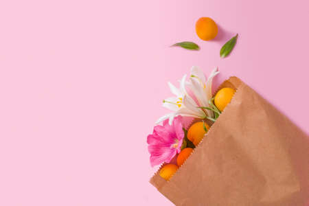 Ripe apricots and lily flowers in a kraft paper bag on a pink background. Waste-free shopping concept, reusable eco-friendly packagingの写真素材