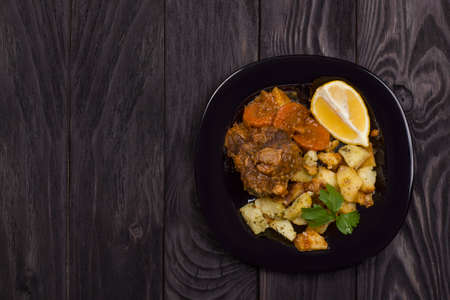 Braised beef tails with vegetables and potatoes on a black plate on a dark wooden background with copy space.の写真素材