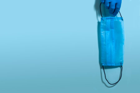 Female hand in a blue sterile rubber glove holding a protective surgical mask on a blue background...