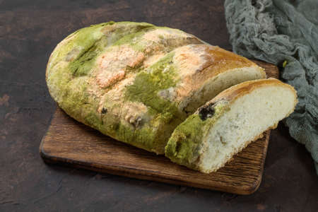 Loaf of moldy white bread on wooden board against dark background.