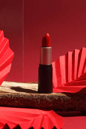 Red lipstick on wooden podium with paper craft and glass on red background. Creative concept photo of cosmetics with hard shadowの写真素材