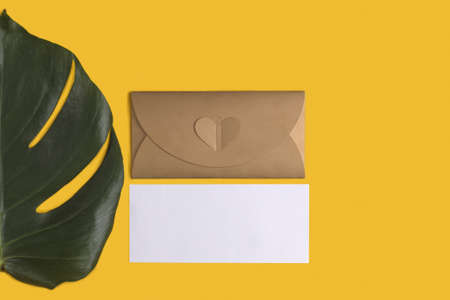 Flat lay with empty greeting card with envelope and monstera leaf on yellow background, top view, copy space. Template for branding and advertisingの写真素材